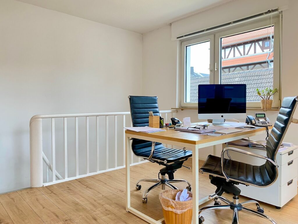 Modernes Büro mit zwei schwarzen Bürostühlen, Holztisch, iMac-Computer und Fenster mit Blick auf ein Fachwerkhaus.