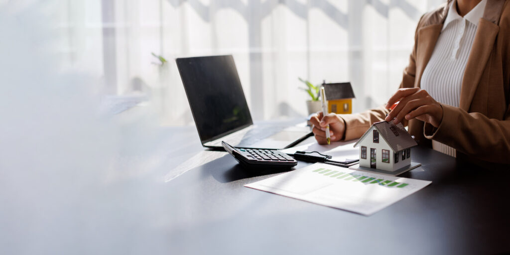 Immobilienmaklerin am Schreibtisch mit Laptop, Taschenrechner und Modellhaus, analysiert Immobilienmarkt und erstellt Finanzplanung.